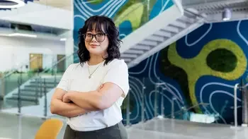 Student facing the camera with arms crossed in front of a staircase inside a campus building