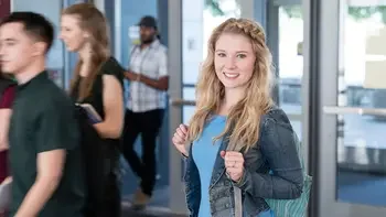 student wearing a backpack smiling at the camera, other students walking into a campus building behind