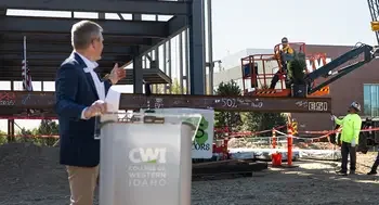 A man at a podium signals to raise a metal construction beam on a work site.