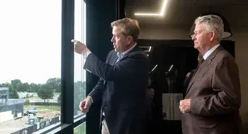 Two men look out the window of a building, one pointing out construction to the other.