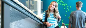 student on a staircase inside a campus building smiling at the camera with students walking down the stairs around her