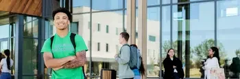 Student wearing a backpack standing outside a campus building with groups of students around him having conversations amongst one another