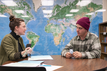 Two people sitting at a table talking in front of a world map